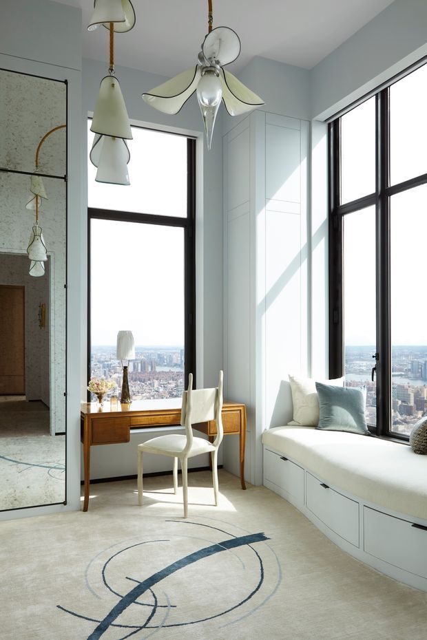 Bright, refined high-rise bedroom or sitting room with floor-to-ceiling corner glazing and expansive city views. A built-in window seat with drawers occupies the right side, accompanied by a white chair, timber writing desk, and sculptural pendant lights. The room has pale blue-grey walls, dark window frames, light carpet, and a large abstract circular-pattern rug. A mirrored or reflective sliding door is visible at left.