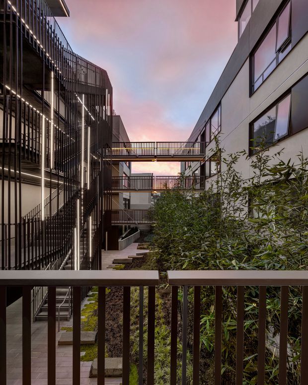A dusk view through a dark metal balustrade into a landscaped courtyard between contemporary multi-storey buildings. The architecture features rectilinear forms, extensive glazing, dark vertical metal screens, illuminated linear lighting, and elevated pedestrian bridges linking the buildings. Dense planting and stepped paved paths occupy the courtyard, with a pink-blue evening sky visible above.