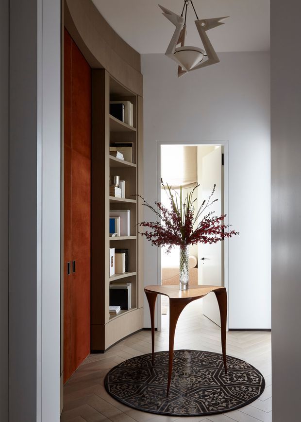 A refined residential entry hall with a curved built-in timber storage wall and open shelving, a narrow sculptural console table, patterned round rug, decorative vase and flowers, and a distinctive geometric pendant light. A doorway opens to a bright adjoining room with pale herringbone flooring. The space combines warm reddish timber, neutral walls, and dark accents in a polished, elegant composition.