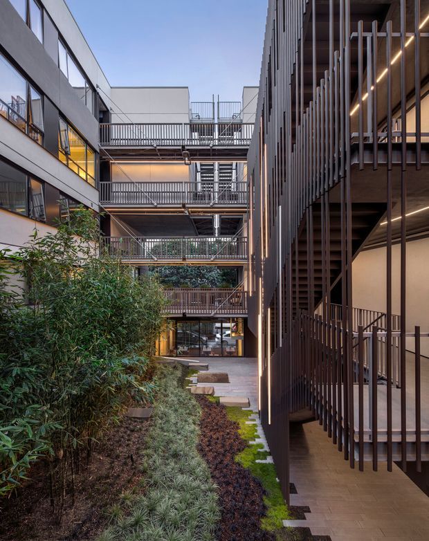 A vertically framed view through a landscaped courtyard within a multi-storey contemporary building complex. Open-air circulation decks with metal balustrades span between pale rendered or concrete-clad wings, while the right-hand facade uses dense vertical metal screening and warm linear lighting. Layered planting, stepping stones, and ground-level glazing create a refined urban courtyard atmosphere.