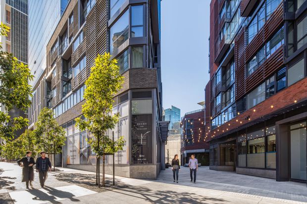A bright urban pedestrian laneway framed by multi-storey contemporary apartment and commercial buildings. The facades combine brick, dark metal-framed glazing, projecting balconies, horizontal screening and ground-level shopfronts. Street trees, paved walkways and festoon lighting create a lively mixed-use public realm.