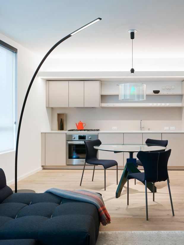 A bright, compact open-plan kitchen and dining area viewed from the living room. The space features pale flat-panel cabinetry, integrated appliances, a light timber floor, a round glass dining table with dark upholstered chairs, a sculptural black arc floor lamp, and a dark blue sofa in the foreground. A large window provides natural light, while a suspended glass pendant hangs above the dining table.