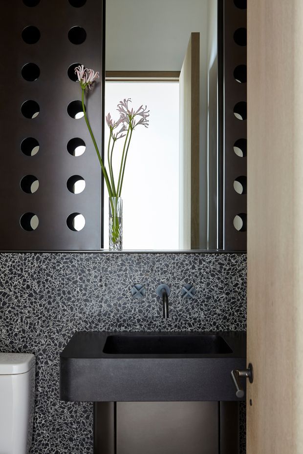 Compact contemporary bathroom or powder room with a dark rectangular basin, wall-mounted black tapware, and a dramatic terrazzo-style grey and white wall finish. A large mirror with a perforated dark panel detail reflects the bright space, while a timber door and partial toilet are visible.