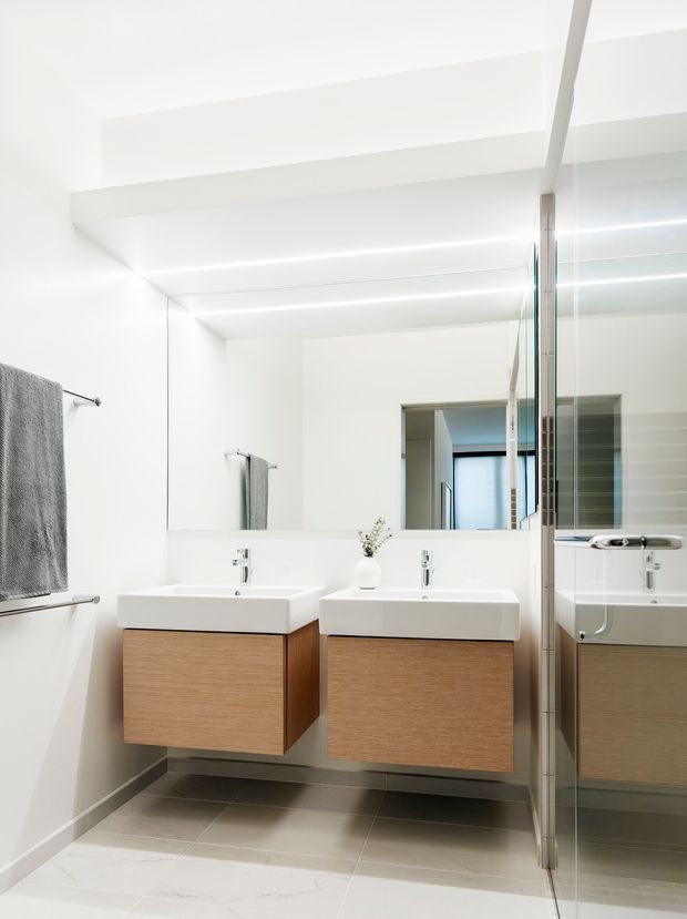 Bright contemporary bathroom featuring twin wall-mounted white rectangular basins with light timber vanity cabinetry, a large mirrored wall, grey tiled flooring, and a glass-enclosed bathtub or shower area. The space has clean white surfaces, integrated linear lighting, and a minimalist built-in design.