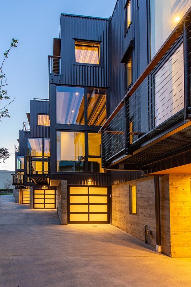 Vertical dusk exterior view of a contemporary multi-level residential development with strongly projecting box-like volumes, dark vertically ribbed metal cladding, extensive glazing, balconies with cable railings, concrete walls, and illuminated sectional garage doors. The camera is positioned in a shared driveway or access court, looking along several attached or closely spaced dwellings. Warm interior and exterior lighting contrasts with the cool blue evening sky.