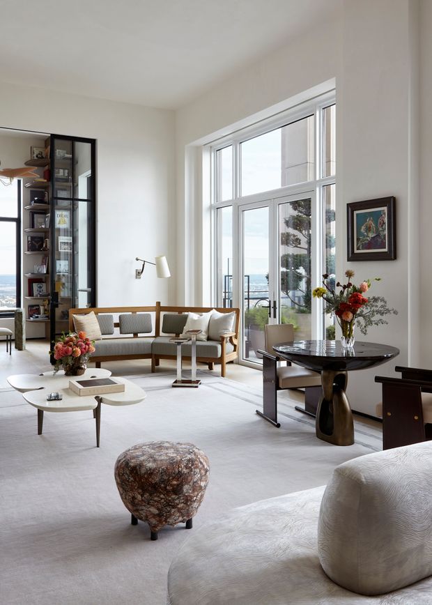 A bright, refined living room with floor-to-ceiling windows and glass doors overlooking a coastal or waterfront setting. The room features a light neutral palette, pale carpet, a tailored upholstered sofa, sculptural coffee tables, a dark sculptural dining table, built-in glass-front shelving, curated artwork, and floral arrangements. Natural light and large glazing create an airy, elegant atmosphere.