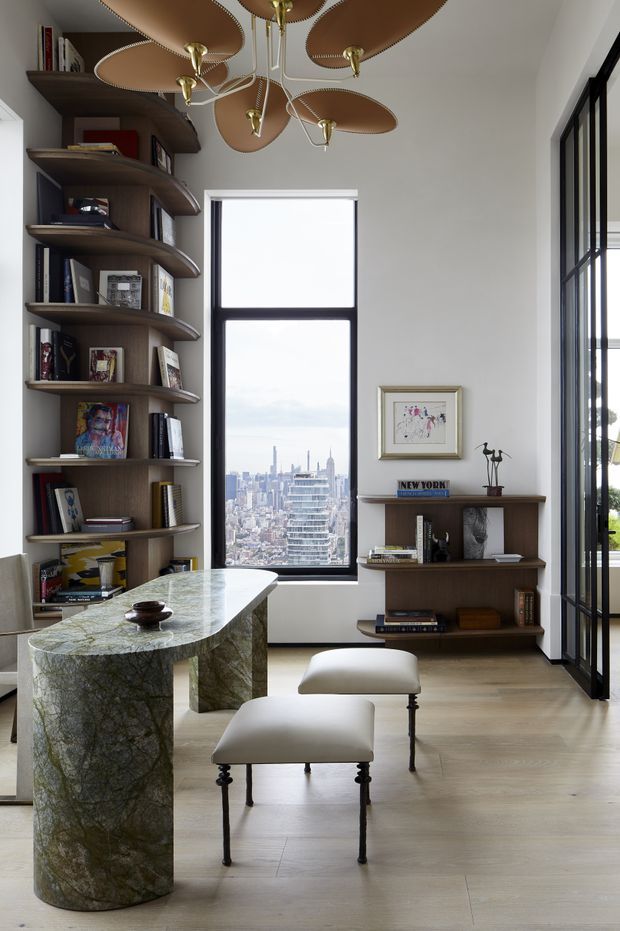 A sophisticated, light-filled interior corner with a sculptural stone console or table in the foreground, two upholstered stools, and extensive custom curved dark-wood shelving displaying books and objects. A large black-framed window provides an urban skyline view, while a glazed black-framed opening is visible on the right. The room features pale timber flooring, white walls, a distinctive brass-and-brown organic pendant light, and curated artwork and decor.