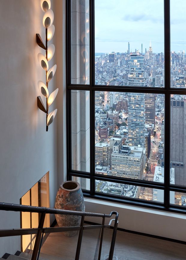 A tall, refined stair landing or stairway with expansive floor-to-ceiling black-framed windows overlooking a dense urban skyline. The interior has pale smooth walls, warm wood flooring, dark metal stair railings, and a sculptural vertical wall sconce with leaf-like illuminated forms. A large rustic ceramic vessel sits near the landing, adding an organic accent to the otherwise sleek, contemporary space.