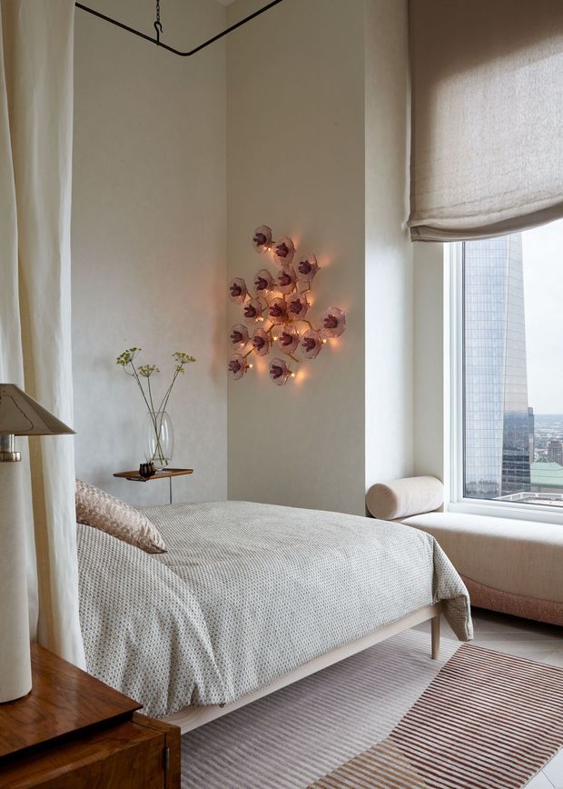 A refined bedroom with a high ceiling, soft warm-neutral plaster walls, and a large city-facing window with a Roman blind. The room features a low upholstered bed dressed in textured white bedding, a sculptural floral wall light, a small minimalist side table with a vase, a timber bedside cabinet, and a striped area rug. Natural light and warm lamp light create an elegant, serene atmosphere.