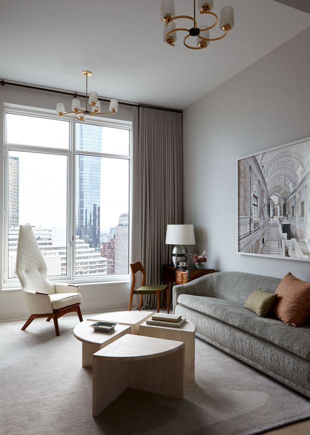 A refined living room in a high-rise setting, with expansive city-facing windows, pale walls, full-height taupe curtains, a textured grey sofa, sculptural light-toned coffee tables, a cream mid-century armchair, and a small timber side table. Gold-toned contemporary chandeliers, a table lamp, and a large architectural corridor artwork add detail. The room has a calm, sophisticated, softly neutral palette.