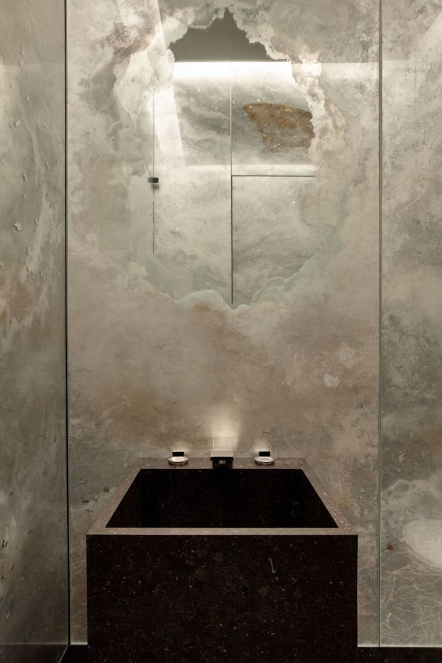 A compact, moody bathroom vanity viewed straight on. The room is clad in large cloudy grey-beige stone slabs with a dramatic irregular mirrored feature panel above the basin. A black stone rectangular basin and matching thick vanity surround sit below a minimalist wall-mounted tap set, with concealed warm lighting reflected in the mirror.