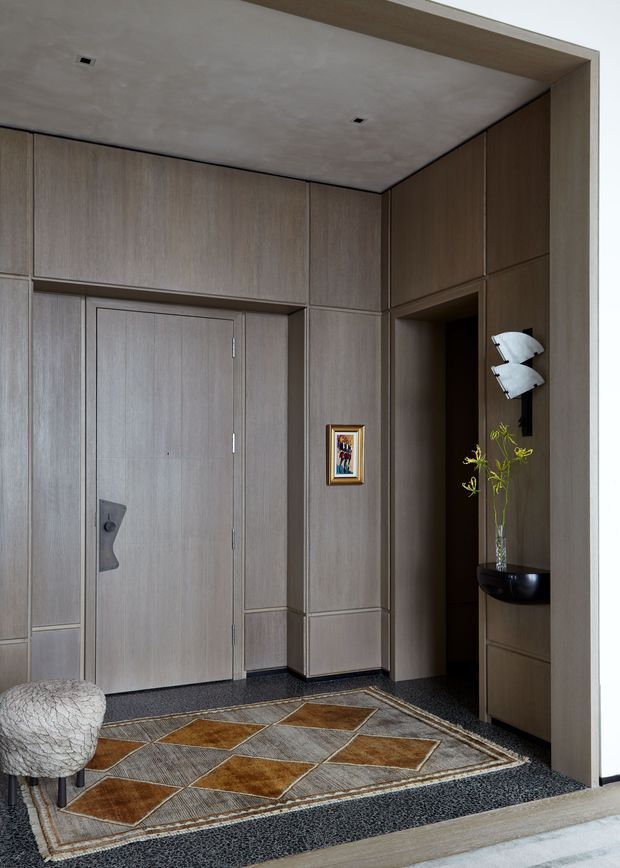Refined interior entryway with floor-to-ceiling grey-brown timber wall panelling and integrated cabinetry surrounding a flush timber door. A recessed niche contains a sculptural wall light, dark vessel and delicate branch arrangement, while a geometric neutral and ochre rug defines the dark textured floor. The space has a minimalist, tailored appearance with concealed recessed lighting and a small upholstered stool.