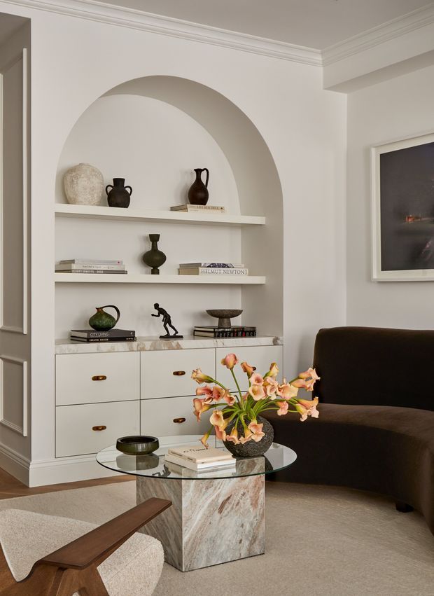 A refined living room with a softly neutral palette, featuring a large arched built-in shelving niche with cream cabinetry and brass pulls. The shelves display books and sculptural ceramics, while a dark curved sofa, glass coffee table, marble pedestal base, upholstered lounge chair, framed artwork, and floral arrangement create a warm, elegant interior.