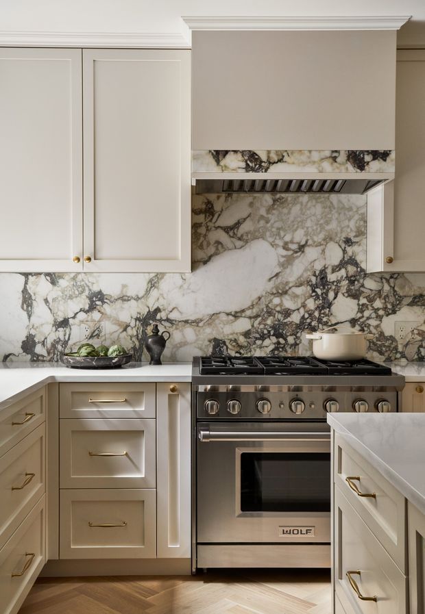 A refined kitchen featuring warm off-white raised-panel cabinetry with brass pulls, a dramatic veined natural-stone backsplash and benchtop, a stainless-steel freestanding range, and a timber herringbone floor. The composition includes a concealed rangehood surround with classic crown moulding and a foreground island.