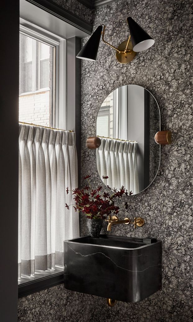 A compact, highly stylised powder-room vanity area with a black marble or stone wall-mounted basin, brass wall-mounted tapware, oval mirror, and a sculptural brass-and-black wall sconce. The walls are covered in a dramatic dark patterned stone-like finish, while a tall window has sheer pleated café curtains with a darker lower band. The palette is dark, monochromatic, and luxurious with brass and burgundy floral accents.