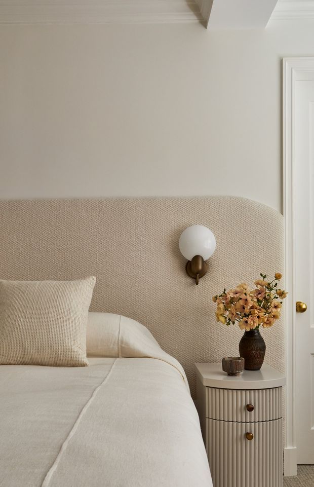 A close-up view of a softly styled bedroom with a curved upholstered headboard in a warm beige textured fabric, layered cream bedding, and a neutral knitted cushion. A spherical wall sconce with a brass-toned mount is fixed to the headboard. Beside the bed is a small fluted white bedside cabinet with brass knobs, topped with a dark vase of dried flowers and a small glass. The room has warm off-white walls, white decorative door trim, and subtle ceiling moulding, creating a calm, refined atmosphere.