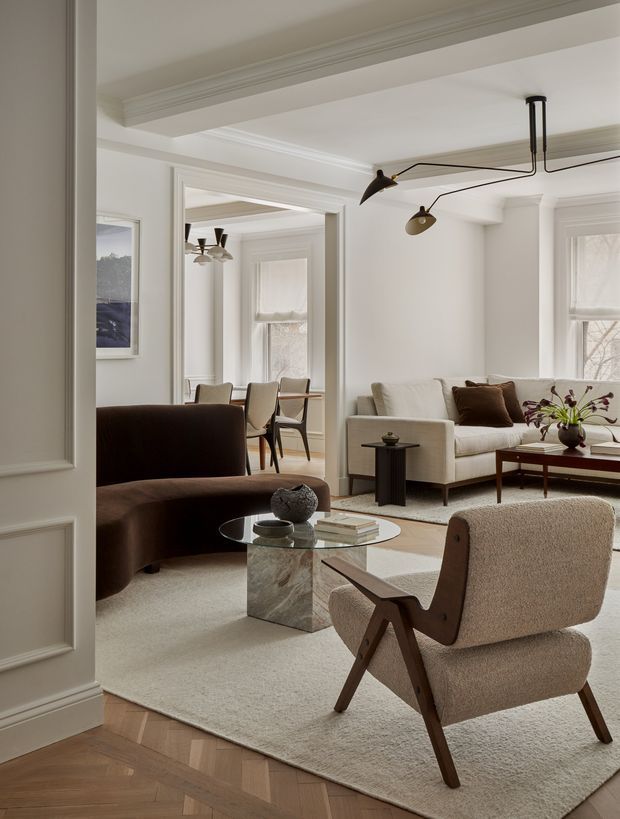 A refined, light-filled living room with a connected dining area visible through a wide interior opening. The room features white paneled walls and decorative ceiling mouldings, a cream sofa, dark brown curved sofa, textured boucle lounge chair with timber arms, glass coffee table with a stone pedestal, and a slim contemporary black pendant light. Pale carpeting contrasts with a warm herringbone timber floor at the entry.