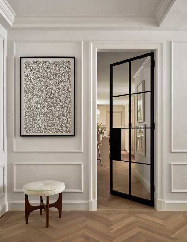 Elegant residential hallway or entry passage with white decorative wall panelling, layered crown moulding and trim, a black-framed glazed internal door, and warm herringbone timber flooring. A small upholstered stool and framed artwork sit against the wall. Through the door, a refined adjoining interior with matching timber flooring and cabinetry is visible.