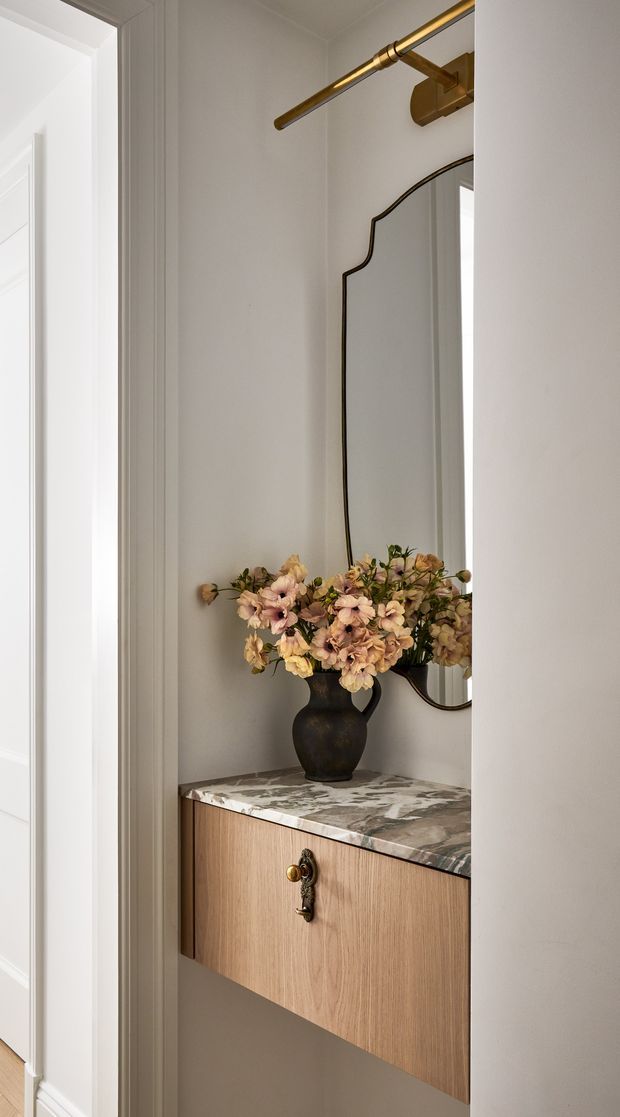 A narrow, elegant entryway vignette featuring a floating timber console with a richly veined stone benchtop, sculptural dark vase and flowers, a decorative framed mirror, and a brass picture light. The surrounding walls and detailed door trim are painted a soft warm white, creating a refined, classic interior.