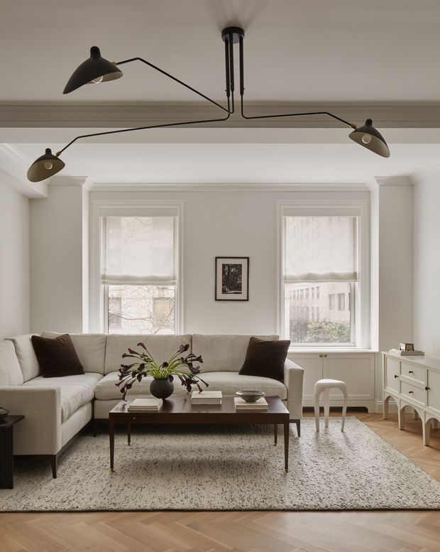 A bright, refined living room with a large pale sectional sofa, dark wood rectangular coffee table, patterned area rug, and a white sideboard. Two tall windows with simple Roman shades are framed by decorative wall mouldings, while a sculptural black multi-arm ceiling light provides a contemporary contrast. Herringbone timber flooring and neutral upholstery create a calm, sophisticated interior.