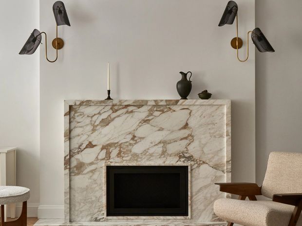 A refined living room vignette centred on a large veined natural-stone fireplace surround with a black firebox. The pale neutral walls and trim are complemented by sculptural dark wall sconces, a small black mantel vessel, and a textured upholstered mid-century-style lounge chair with a timber arm. The composition is symmetrical, calm, and understated, with a strong marble fireplace feature.