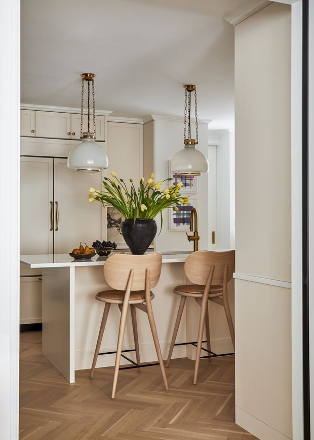 A refined, warm-toned kitchen viewed from an adjacent opening. The space features cream painted shaker-style cabinetry, a pale stone island with integrated seating, sculptural timber bar stools, brass hardware and tapware, white glass pendant lights suspended from chains, and a dark statement vase with yellow flowers. Herringbone timber flooring and traditional wall mouldings add character, while the overall styling is elegant and contemporary-classic.