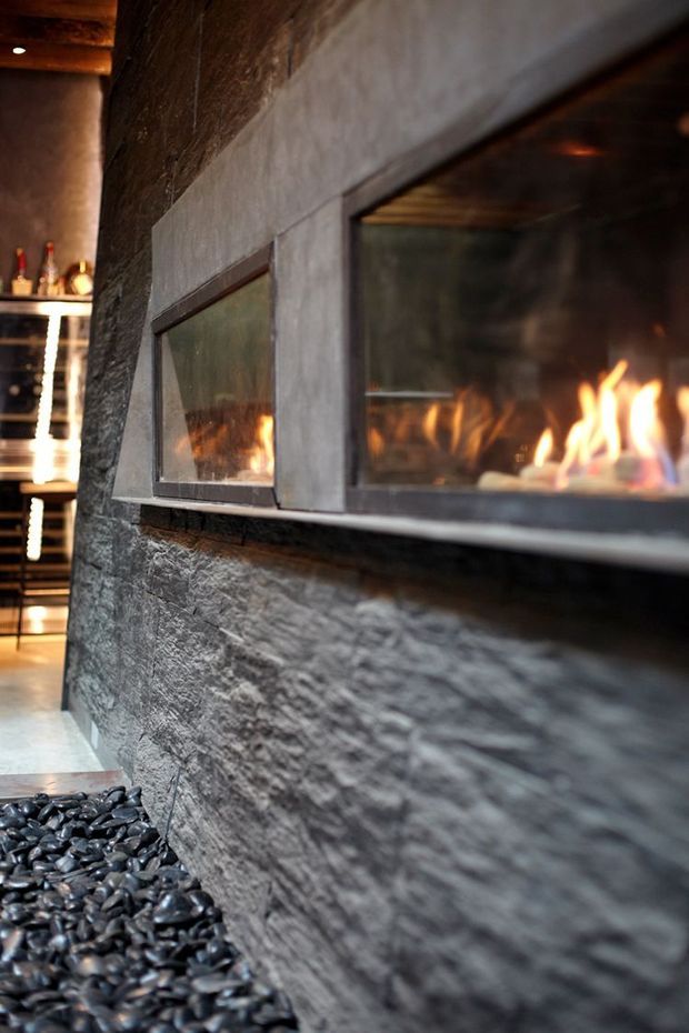 Close-up interior architectural detail of a large built-in fireplace with visible flames behind dark metal-framed glass panels. The fireplace is set into a heavily textured dark grey stone wall, with a bed of smooth black decorative pebbles at its base. A warmly lit bar or shelving area is partially visible in the background.