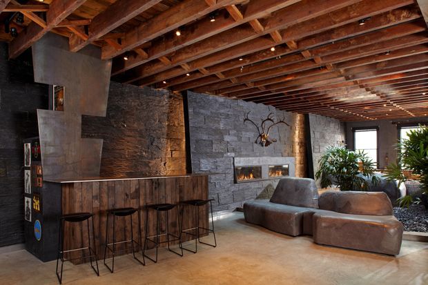 Rustic industrial-style living area with an exposed timber beam ceiling, rough stone feature walls, a large integrated fireplace, and a dark timber bar counter with stools. The room includes low, sculptural brown seating, track lighting, indoor planting, and large windows, creating a warm, textured lounge atmosphere.