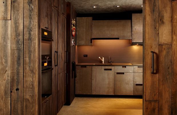 Compact, moody contemporary kitchen with extensive dark timber cabinetry and wall panelling. Integrated appliances are concealed within tall joinery on the left, while a timber-fronted island or lower cabinet run faces a warm metallic-toned splashback and under-cabinet lighting. Black handles, recessed downlights, and a textured dark ceiling add contrast and a refined rustic-industrial character.