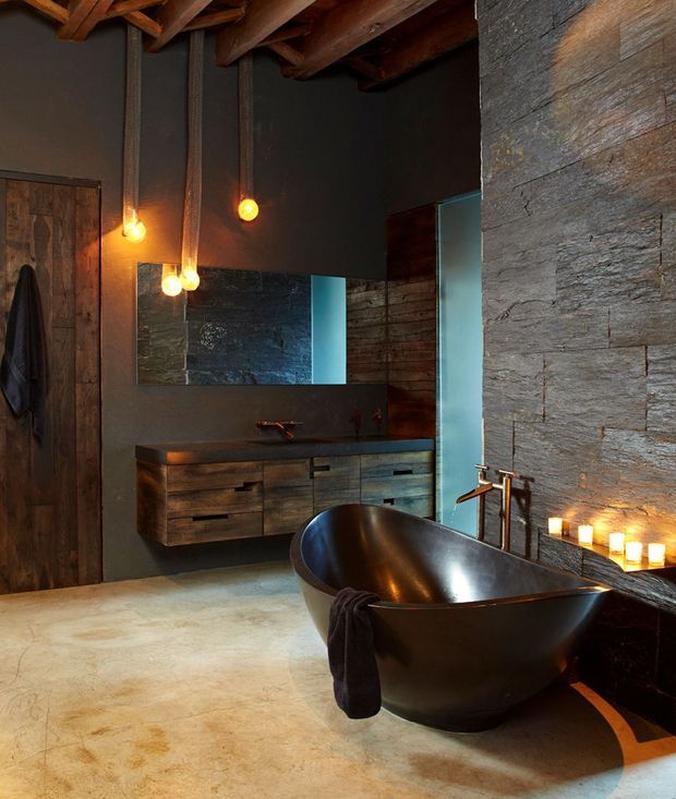 A dramatic rustic bathroom featuring a freestanding dark bathtub, wall-mounted timber vanity with integrated basin, large horizontal mirror, exposed timber ceiling beams, dark textured wall surfaces, and warm pendant and candle lighting. The room combines raw natural materials with a moody, spa-like atmosphere.