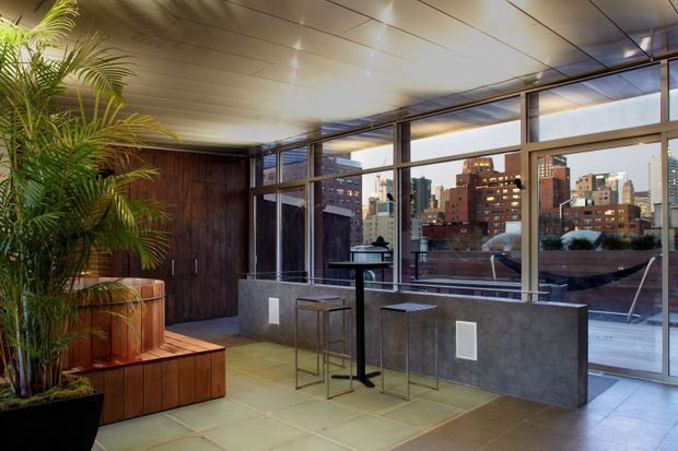 A contemporary rooftop or upper-level interior lounge with expansive floor-to-ceiling glazing overlooking an urban skyline. The space features a large tropical planter, warm timber wall cladding and built-in seating, a polished concrete or stone-height counter, slim metal bar stools and tables, and a light panelled ceiling. The atmosphere is refined, urban and warmly lit.
