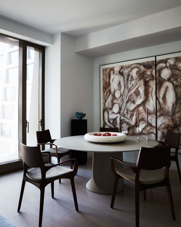 Refined dining room with a round pedestal dining table surrounded by dark wood armchairs, set against a large abstract artwork. Full-height windows with dark timber frames bring in abundant daylight, while the restrained palette and sculptural furniture create an elegant contemporary interior.