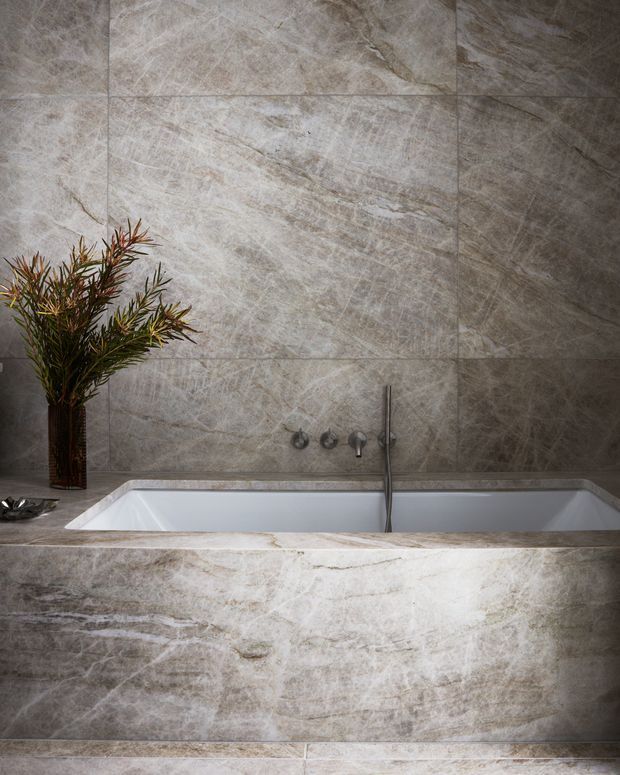 A luxurious bathroom featuring a built-in rectangular bathtub clad in veined grey-beige stone, matching large-format stone wall and floor tiles, and minimalist dark metal wall-mounted tapware. A tall vase with foliage provides a natural decorative accent.
