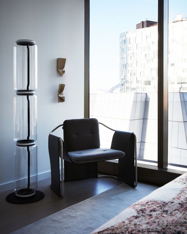 A contemporary interior corner with a black leather or upholstered armchair beside floor-to-ceiling windows. The room has dark timber flooring, a pale wall, a sculptural black floor lamp, and small wall-mounted decorative fixtures. A patterned rug enters the foreground, while a bright modern building is visible outside.