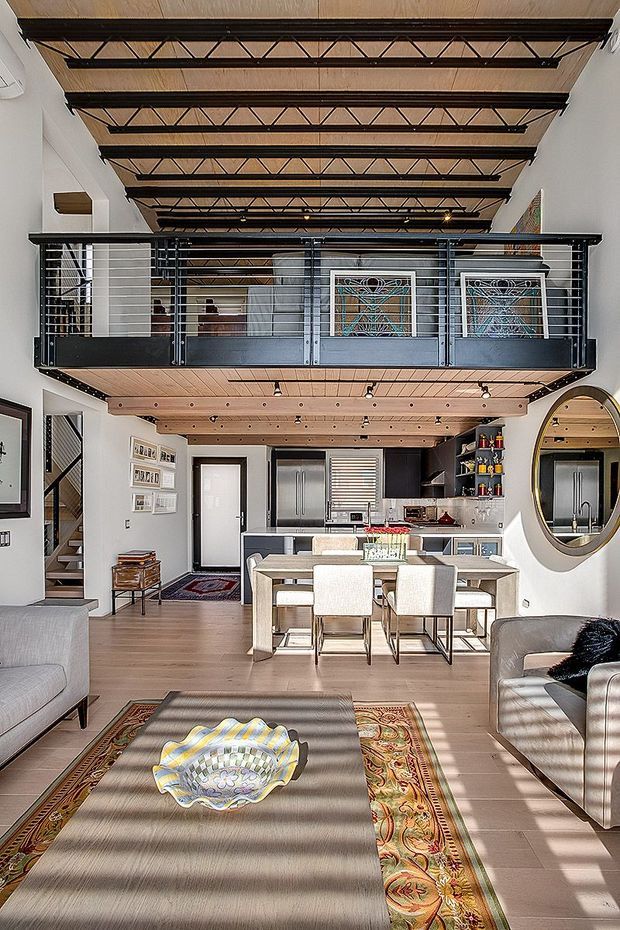Bright open-plan loft interior combining a living room, dining area and kitchen beneath a high exposed timber-and-steel truss ceiling. A black metal mezzanine balcony spans the upper level, with decorative glazed doors behind it. The main level features pale timber flooring, a dining table with upholstered chairs, a contemporary kitchen with dark cabinetry and stainless-steel appliances, a large round wall mirror, and a patterned area rug. The space has strong industrial structural detailing softened by warm timber finishes and traditional furnishings.