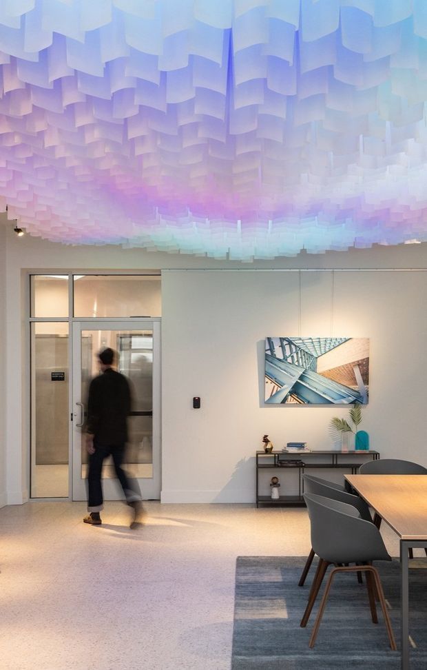 A contemporary commercial interior, likely an office or meeting space, with a pale neutral palette and a highly distinctive suspended ceiling installation made of layered translucent rectangular panels illuminated in soft blue, lavender, pink and mint tones. A glazed framed door opens to another room on the left, while a console table, framed artwork, dining or meeting table, grey upholstered chairs and a large muted rug occupy the space. The room has a polished, refined and softly atmospheric character.