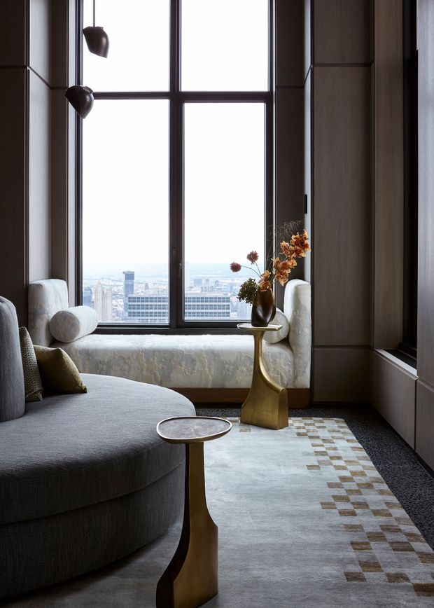A refined high-rise living room with a large dark-framed grid window overlooking a city skyline. The room features a curved grey sectional sofa, a deep window seat with light upholstery, dark wood wall panelling and built-in cabinetry, sculptural bronze-gold side tables, a floral arrangement, and a pale area rug with a geometric checkered border. The mood is sophisticated, warm, and quietly luxurious.