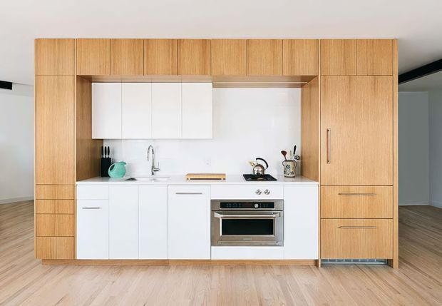 Compact contemporary kitchen with a white fitted cooking run framed by full-height light timber cabinetry. The kitchen includes white lower and upper cabinets, a built-in stainless-steel oven and cooktop, sink, and open worktop, with integrated storage towers and drawers on both sides. Light timber flooring and a simple white ceiling create a bright, clean, minimalist appearance.