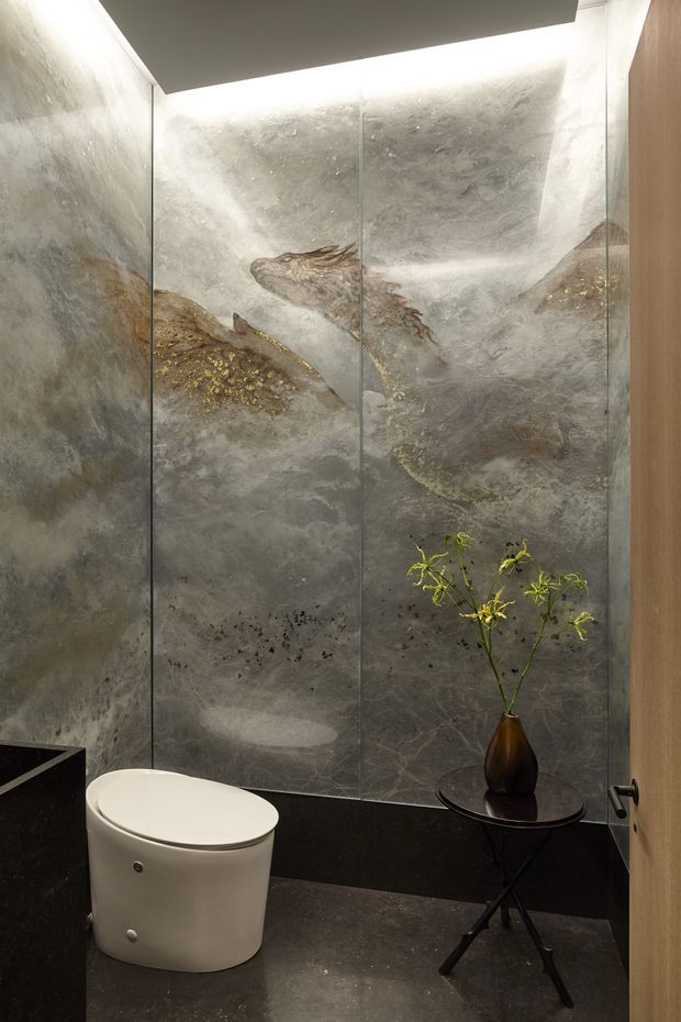 A compact, refined bathroom with a white modern toilet, dark flooring and lower wall trim, and dramatic grey veined stone-look wall panels. A large illuminated feature wall creates a sculptural backdrop, complemented by a small black side table with a vase and a timber door.