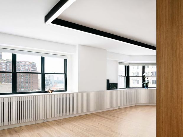 Bright, mostly empty contemporary interior with pale timber flooring, white walls and ceiling, expansive black-framed windows, and a long vertical slatted radiator or built-in wall treatment beneath the glazing. A linear black ceiling detail adds contrast, while a timber-clad vertical element frames the right side. Urban apartment buildings are visible outside.