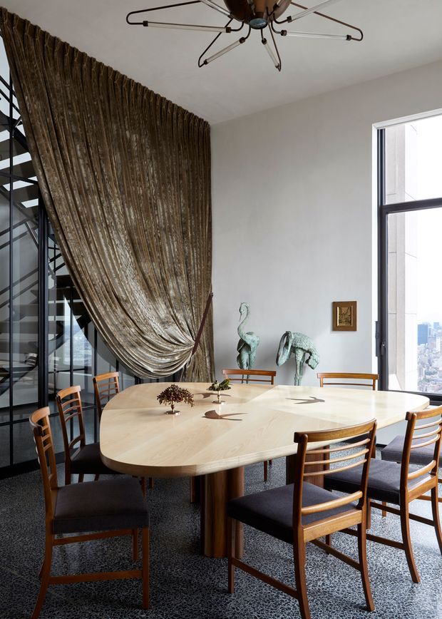A refined dining room with a large rounded rectangular light timber dining table surrounded by mid-century style wooden chairs with dark upholstered seats. A dramatic floor-to-ceiling bronze-brown drapery wall frames a black steel and glass staircase, while a large window provides an urban city view. The room features a sculptural radial ceiling light, pale walls, patterned dark carpet, and decorative bird sculptures on a console.