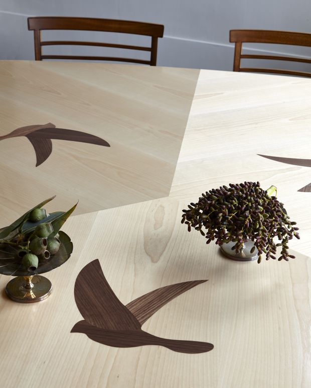 Close-up view of a light timber dining table with geometric panel joints and dark wood inlay motifs resembling flying birds. Two dark timber dining chairs are visible behind the table, along with decorative potted plants placed on the tabletop. The image emphasizes the table's crafted surface and contrasting wood detailing.