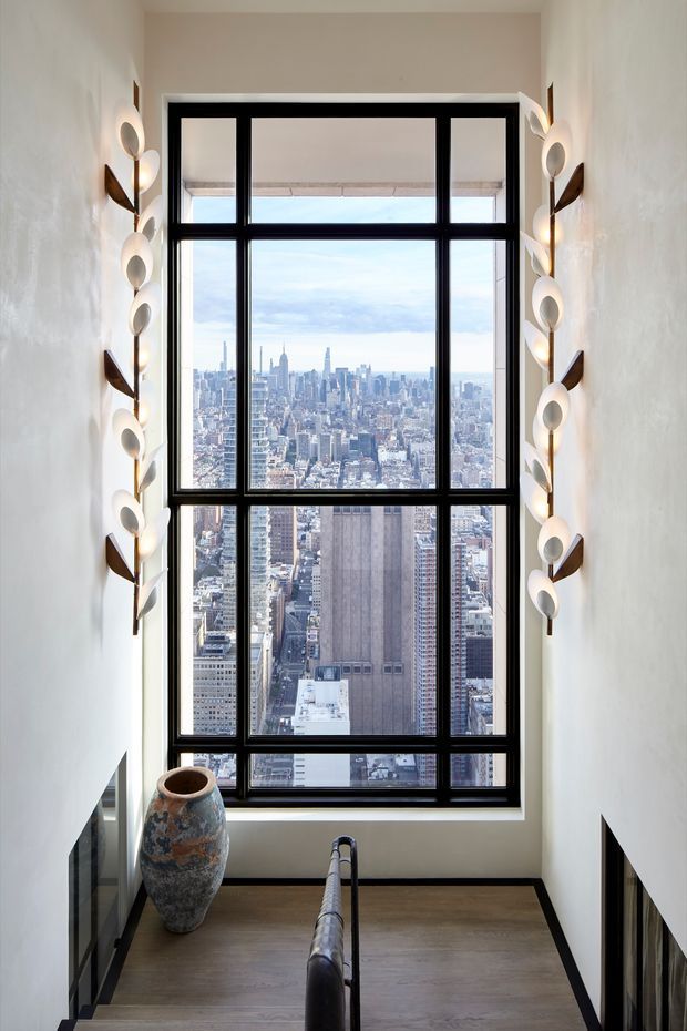 A narrow, tall stair landing or stairway terminates at a large black-framed multi-pane picture window overlooking a dense city skyline. Sculptural wall sconces line both sides of the opening, while dark lower wall panelling, light plaster walls, and a timber-toned floor frame the view. A dark stair handrail rises in the foreground and a large decorative ceramic vessel sits beside the window.