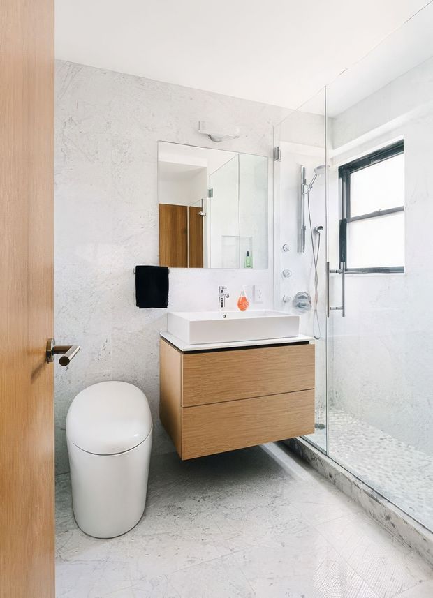 Bright contemporary bathroom with a frameless glass walk-in shower, white vessel-style basin, floating light timber vanity, wall mirror, rounded white toilet, and pale stone-look wall and floor finishes. A timber door and black-framed window add contrast.