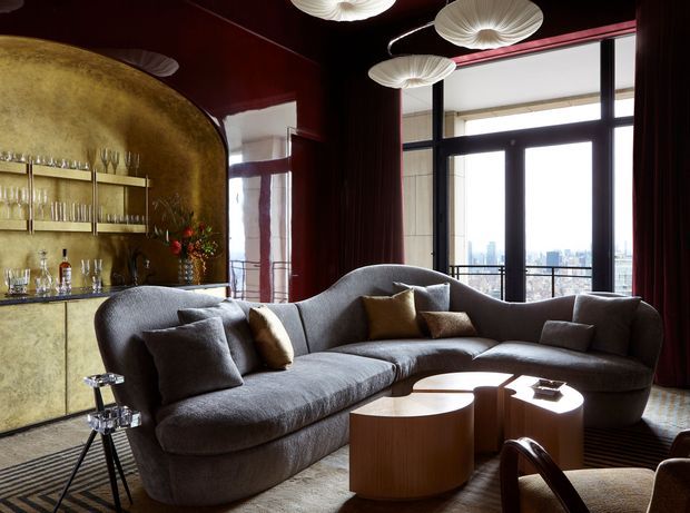 A sophisticated living room with a large curved grey sectional sofa, nested rounded wooden coffee tables, and a dramatic built-in gold-toned bar and display cabinet. Deep burgundy walls and curtains, tall black-framed glazing, sculptural pendant lights, and an urban skyline view create a luxurious, moody interior.
