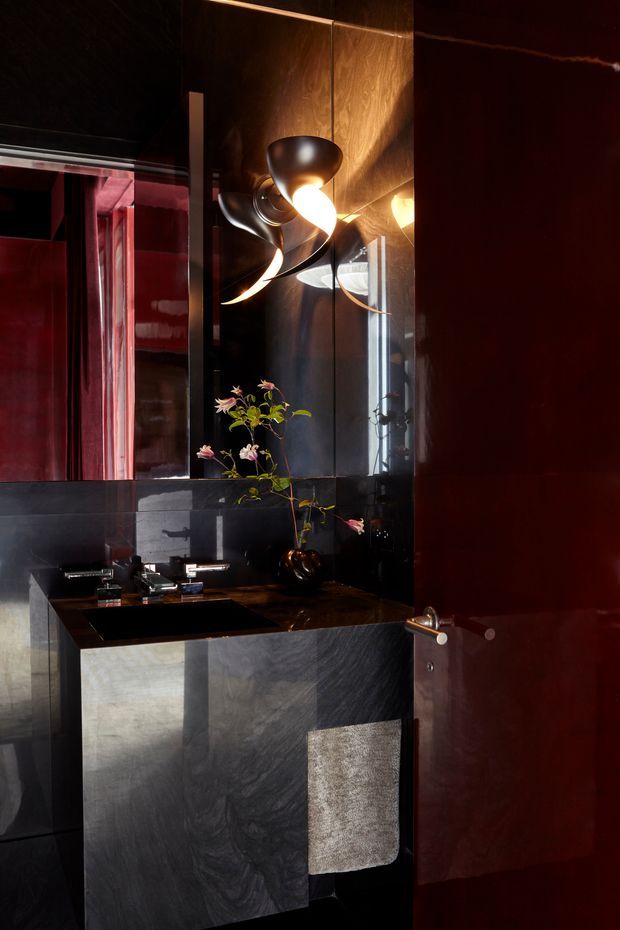 A dramatic, compact bathroom featuring a floating dark stone vanity with integrated rectangular basin, chrome tapware, and a large mirrored wall. Glossy black wall surfaces and deep red timber or lacquered door panels create a moody, high-contrast interior. A sculptural metallic wall light, reflected in the mirror, provides warm illumination, while a small branch arrangement adds a natural accent.