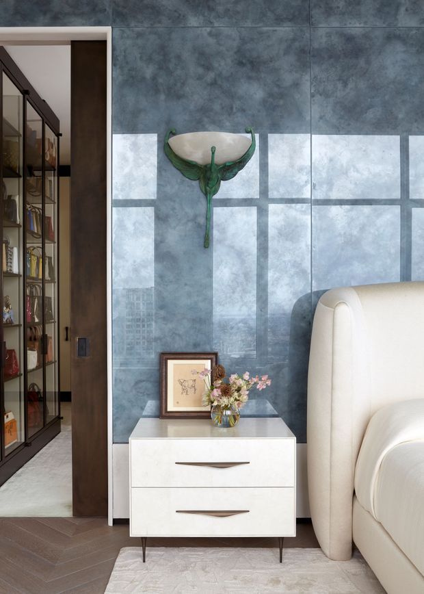 A refined bedroom corner featuring a cream upholstered bed, a white two-drawer bedside table with tapered legs, and a blue-grey marbled feature wall. A decorative sculptural wall piece, framed artwork, and flowers add detail. To the left, a dark-framed glazed built-in shelving unit is visible through an opening. The floor has a dark herringbone timber finish, and bright window reflections create a geometric pattern across the wall.
