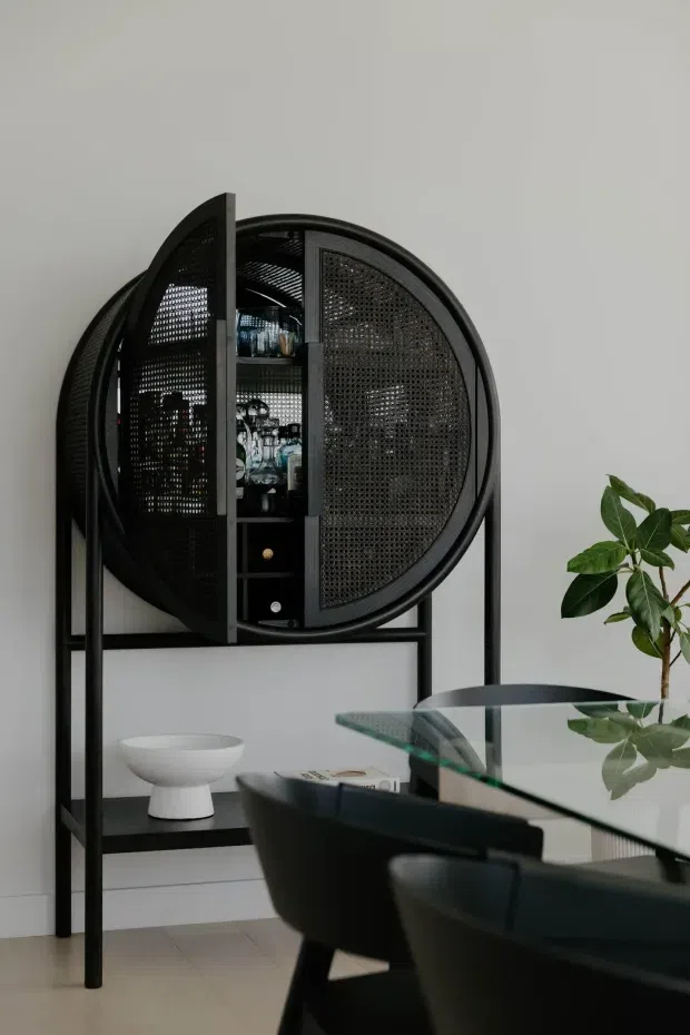 A refined dining area with a striking black circular rattan-front drinks cabinet on a tall black metal frame. A glass dining table with dark upholstered chairs occupies the foreground, while a small sculptural white bowl and a leafy indoor plant add restrained decorative elements. The room has pale walls, light flooring, and a dark, monochromatic furniture palette.