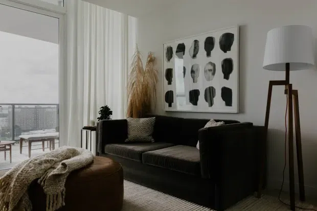 A refined contemporary living room with a black sofa, textured neutral cushions, a round brown leather ottoman, and a tall tripod floor lamp. Floor-to-ceiling sheer curtains frame a balcony and city view, while a large monochrome artwork and dried grasses add visual interest. The room has a calm, muted neutral palette with layered textures and abundant natural light.
