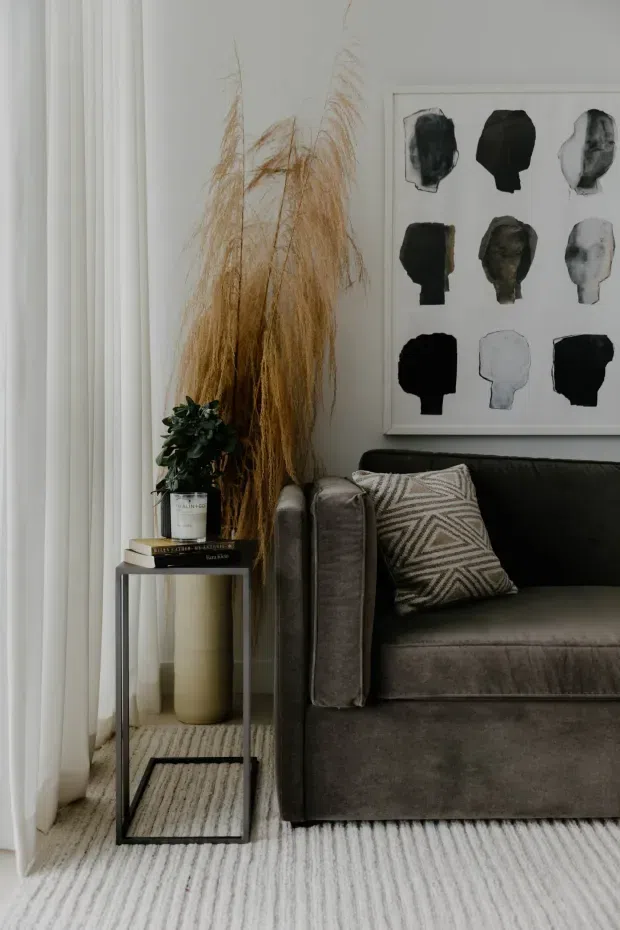 A close-up of a refined living room corner featuring a deep grey upholstered sofa, a geometric neutral cushion, a slim black side table with books and a potted plant, tall dried grasses, sheer white curtains, and a framed abstract artwork. The space has a muted, layered neutral palette and a softly styled contemporary feel.